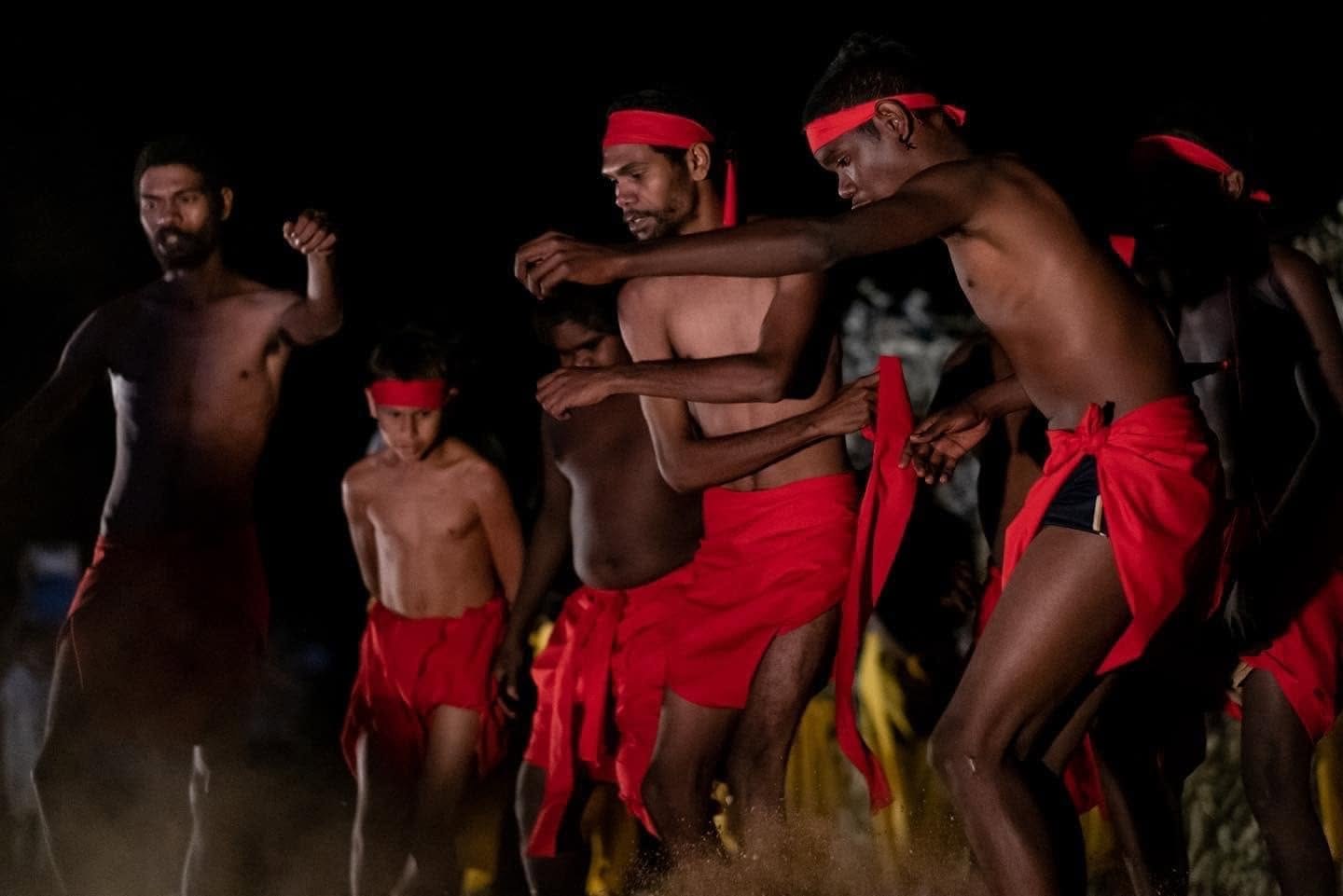 Men and boys in red lap-laps and headbands performing a traditional dance at night