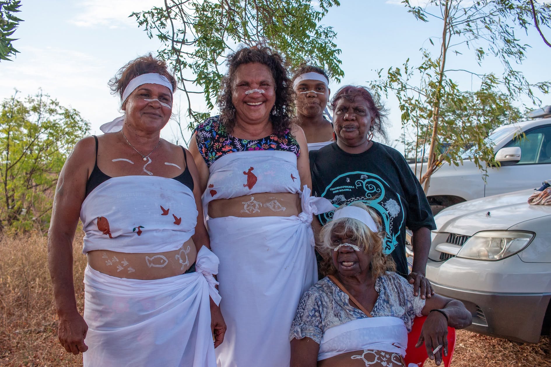 A group of women in white wraps and headbands with painted designs, smiling together outdoors
