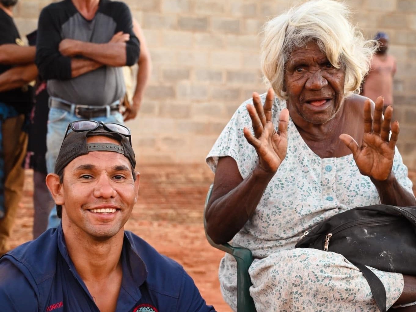 An Elder seated on a chair beside a smiling young man in a ranger uniform on red dirt