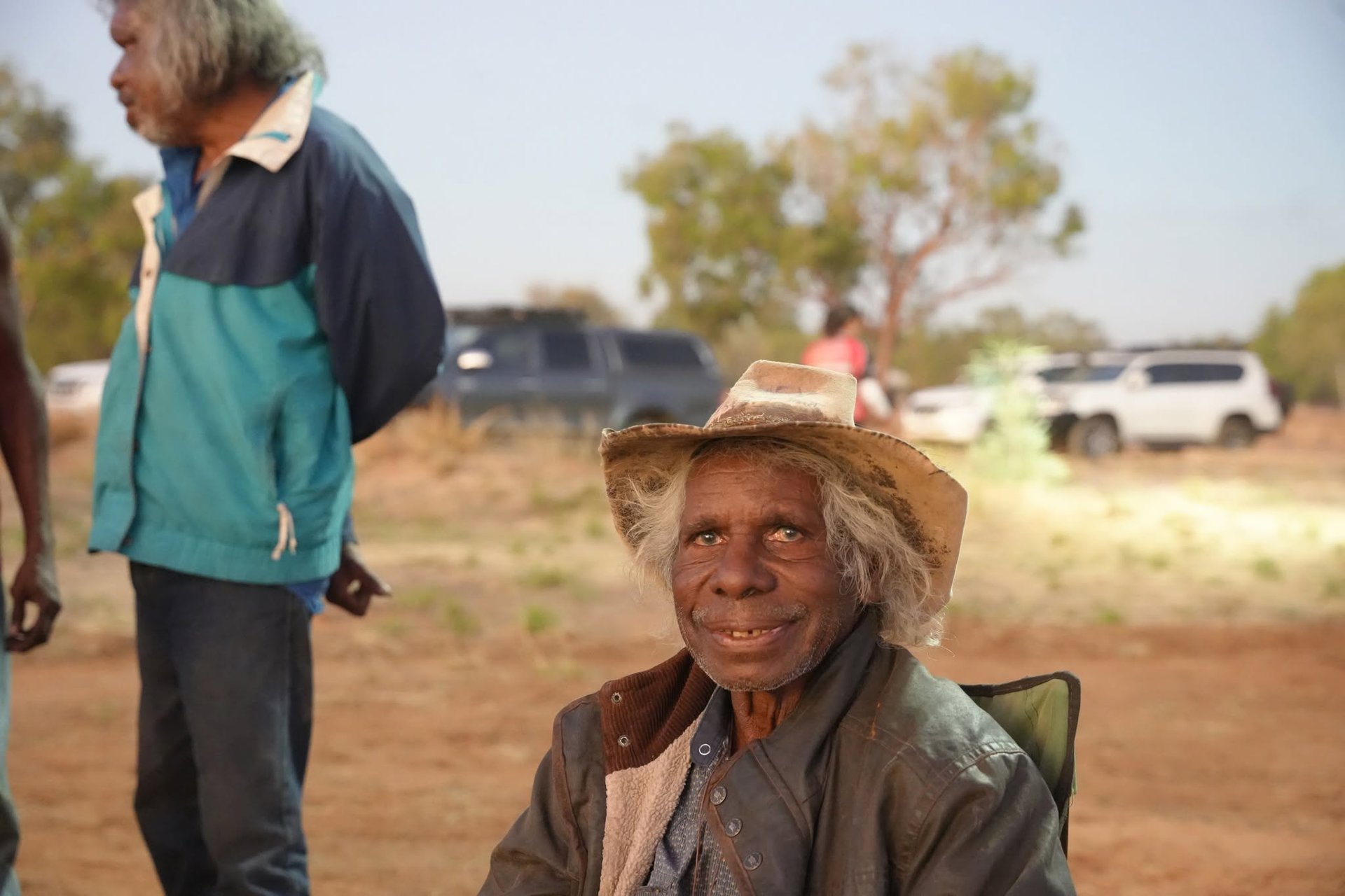 An Elder in a wide-brimmed hat smiling, seated outdoors