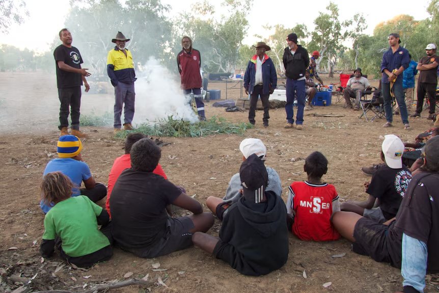 Children sitting on the ground listening to Elders and community leaders beside a smoking fire