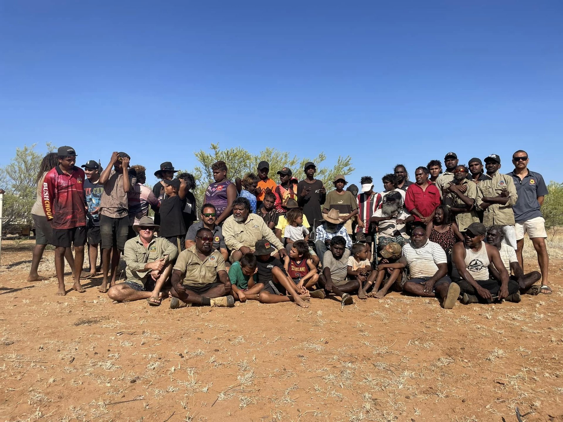 A large group of Elders, young people and families gathered together on red dirt under a blue sky