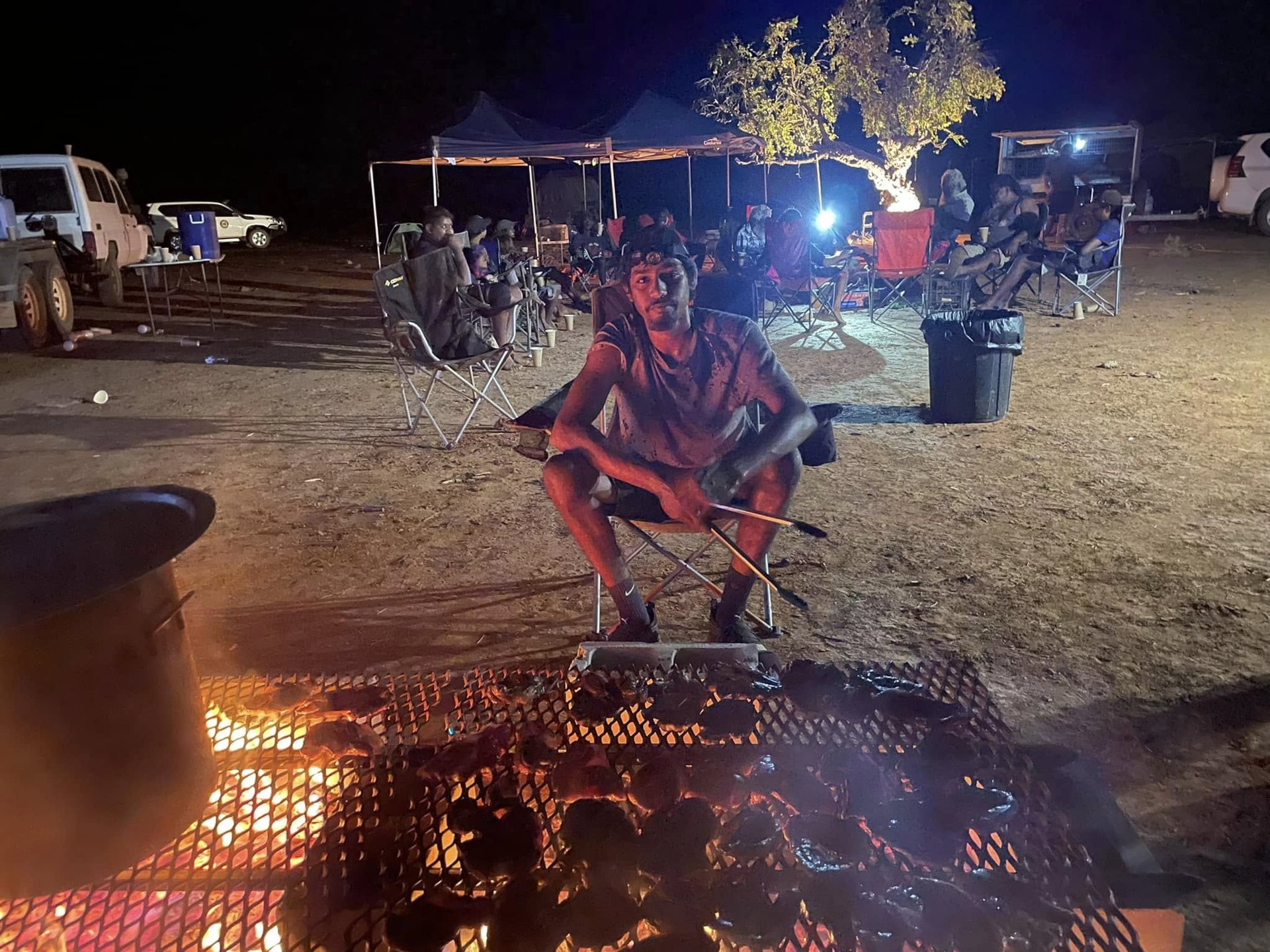A young man sitting beside a campfire grill at a night camp, with tents and vehicles behind